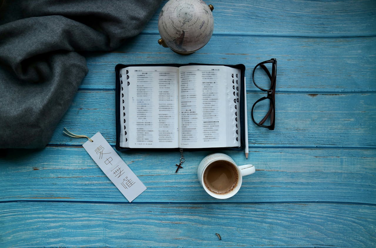 An open Bible with a bookmark, coffee, glasses, and a globe on a blue wooden table.