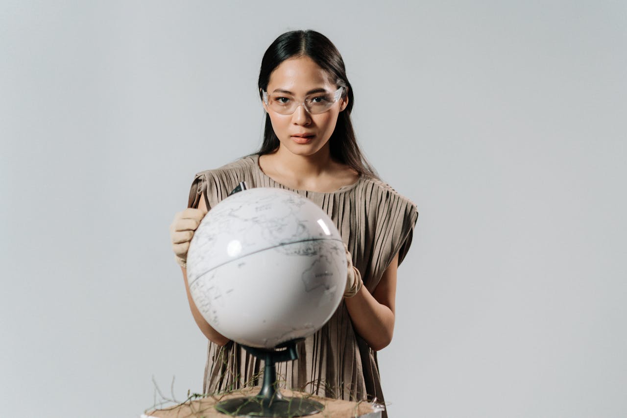 Portrait of an Asian woman in safety glasses holding a globe, set against a neutral background.